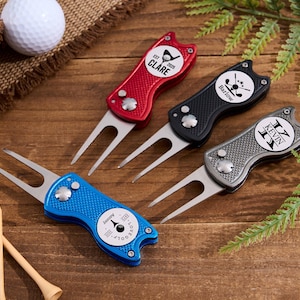 Könnte beinhalten: Vier personalisierte Golf-Divot-Werkzeuge in Rot, Schwarz, Blau und Silber, mit Namen und Initialen. Jedes Werkzeug hat eine Metallgabel und ein kreisförmiges Design mit Text. Ein weißer Golfball und Holztees sind ebenfalls sichtbar.