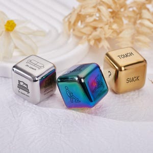 May include: Three metal dice with engraved text and illustrations. One die is silver with "On couch", "In bedroom", and "On balcony". Another is iridescent with a graphic. The third is gold with "TOUCH" and "SUCK".