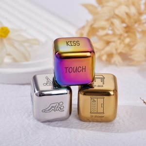 Könnte beinhalten: Drei Metalldürfel mit verschiedenen Designs. Ein Würfel ist irisierend mit den Gravuren "KISS" und "TOUCH". Die anderen beiden Würfel sind silber- und goldfarben, mit Illustrationen und Text wie "on desk" und "in shower".