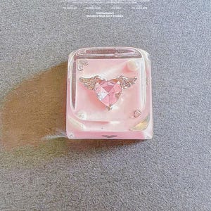 May include: A pink and clear keycap with a silver winged heart charm. The keycap is a popular accessory for mechanical keyboards.