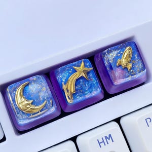 Puede incluir: Tres teclas de teclado personalizadas con diseños temáticos espaciales. Las teclas presentan una luna creciente dorada, una estrella fugaz y un cohete, sobre un fondo de galaxia azul y púrpura. Las teclas están en un teclado blanco.