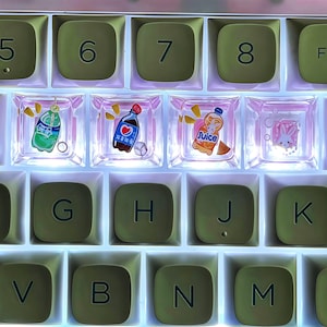 May include: Close-up of a mechanical keyboard with custom keycaps. The keycaps are olive green with white lettering. Some keycaps are clear with colorful illustrations of drinks and a bunny. The keyboard is backlit with a soft glow.