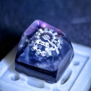 以下が含まれることがあります： 内部に白い歯車のようなデザインが埋め込まれた、半透明の濃い青と紫の樹脂アート作品。 樹脂は小さなピラミッドの形をしており、白い型の中にあります。 この作品は光沢のある仕上がりで、内側から照らされています。