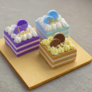 Puede incluir: Tres bizcochos en miniatura con capas coloridas y glaseado blanco sobre una tabla de madera. Los pasteles están decorados con galletas y la palabra "love" o "style" en letras doradas. Los pasteles son morados, azules y beige.