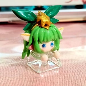 Może przedstawiać: Mała figurka postaci z zielonymi włosami, dużym zielonym nakryciem głowy w kształcie liścia i złotymi akcentami. Postać ma niebieskie oczy, różowe policzki i jest zamontowana na przezroczystej, kwadratowej podstawie. Figurka znajduje się na różowej powierzchni.