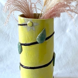 May include: A yellow ceramic vase with a floral design and green leaves. The vase is decorated with black lines and has a textured surface.