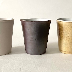 Puede incluir: Tres tazas de cerámica en diferentes colores: blanco, gris oscuro y marrón claro. Las tazas son todas del mismo tamaño y forma, y tienen un diseño simple y moderno.