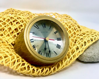 May include: A gold-toned clock with Roman numeral markings and black hands. The clock face is white with black markings for the numbers 13 through 23. The clock face also has the words "SHIPS TIME" printed on it. The clock is sitting on a yellow crocheted surface.