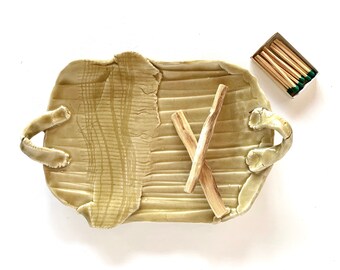May include: A ceramic tray with two wooden sticks and a box of matches. The tray has a textured surface and two handles. The tray is a light beige color.