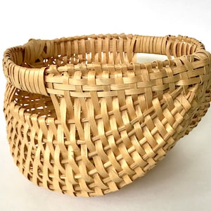May include: A handwoven basket with a natural, light brown color. The basket is made of woven strips of material, creating a textured surface. The basket has a rounded shape with a curved rim and a handle.