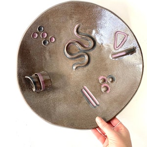 May include: A brown ceramic plate with a raised design of a snake, geometric shapes, and dots in pink and black.