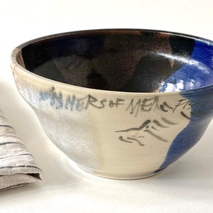 May include: A white ceramic bowl with blue and black glaze. The bowl has a handwritten inscription that reads "Shors of Mesa Fish".