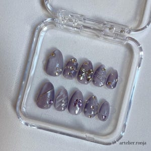 May include: A set of lavender-colored press-on nails in a clear plastic case. Each nail is adorned with silver and gold embellishments, including rhinestones, a bow, and metallic accents. The nails are oval-shaped and feature various designs.