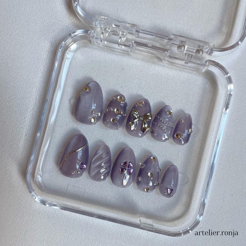 May include: A set of lavender-colored press-on nails in a clear plastic case. Each nail is adorned with silver and gold embellishments, including rhinestones, a bow, and metallic accents. The nails are oval-shaped and feature various designs.