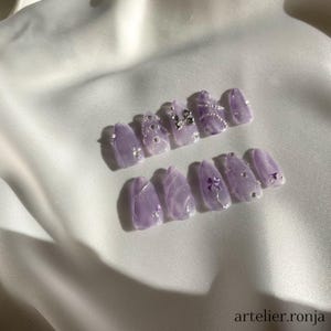 May include: A set of lavender-colored artificial nails with a marble effect. Some nails are adorned with silver rhinestones and small bows. The nails are arranged on a white satin surface. The text "artelier.ronja" is visible.