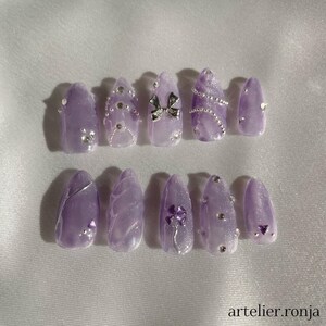May include: A set of ten lavender-colored press-on nails. Each nail is adorned with silver embellishments, including rhinestones, a bow, and a clover design. The nails have a glossy finish and are arranged on a white surface. The text "artelier.ronja" is visible.