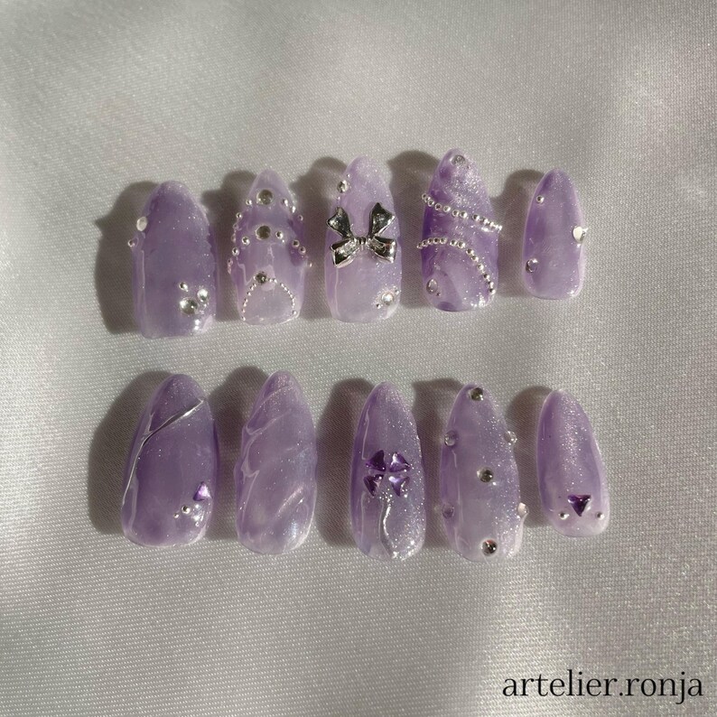May include: A set of ten lavender-colored press-on nails. Each nail is adorned with silver embellishments, including rhinestones, a bow, and a clover design. The nails have a glossy finish and are arranged on a white surface. The text "artelier.ronja" is visible.