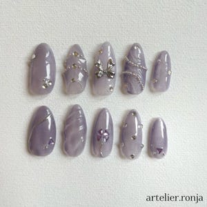 May include: A set of ten lavender-colored artificial nails. Each nail is decorated with silver accents, including rhinestones, chains, and a bow. The nails are oval-shaped and have a glossy finish. The text "artelier.ronja" is visible at the bottom right.