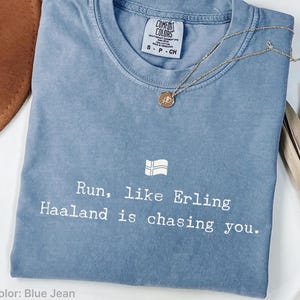 May include: A blue jean coloured t-shirt with a crew neckline and a white graphic print that reads, Run, like Erling Haaland is chasing you, accompanied by a small Norwegian flag icon. The shirt features a Comfort Colors brand label at the neck.