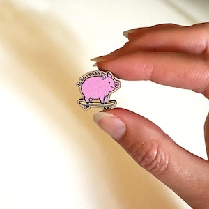 May include: A wooden pin featuring a pink pig on a skateboard. The pig has a smiling face and the words "Go Vegan" are printed above it. The pin is held between fingers.