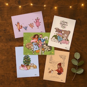 Peut inclure: Plusieurs cartes de vœux illustrées avec des scènes d'animaux et de familles. L'une d'elles porte l'inscription "Happy Mother's Day". D'autres cartes représentent une famille avec des animaux de compagnie, un cochon avec un chapeau de fête et un enfant avec un lapin. Les cartes sont sur une surface en bois.