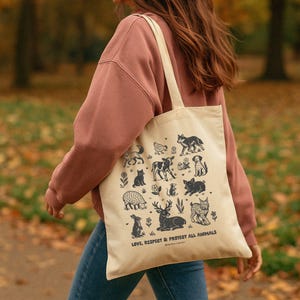 May include: Beige tote bag featuring black illustrations of various animals, including a wolf, cow, cat, and deer. The text "LOVE, RESPECT & PROTECT ALL ANIMALS" is printed at the bottom. The bag has a simple design with long straps.