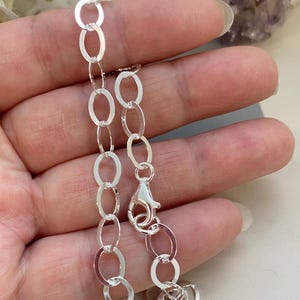 May include: A silver chain bracelet with oval links. The bracelet is made of shiny metal and features a lobster clasp closure. The links are connected in a repeating pattern, creating a delicate and elegant design.