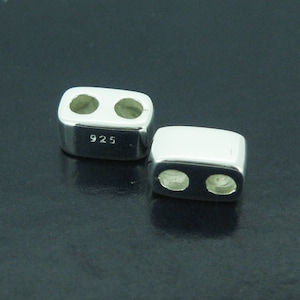 Puede incluir: Dos cuentas rectangulares plateadas brillantes, cada una con dos agujeros. Las cuentas tienen bordes redondeados y el número "925" está estampado en una de ellas. Se presentan sobre un fondo oscuro.