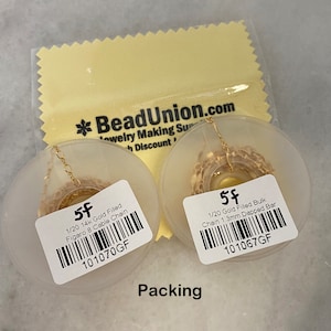 K&ouml;nnte beinhalten: Zwei Spulen mit goldgef&uuml;llter Kette, eine mit der Aufschrift "1/20 14k Gold Filled Figaro 8 Cable Chain 101070GF" und die andere mit der Aufschrift "1/20 Gold Filled Bulk Chain 1.3mm Dapped Bar 101067GF". Beide Spulen befinden sich auf einem wei&szlig;en Hintergrund mit dem Wort "Packing" darunter.