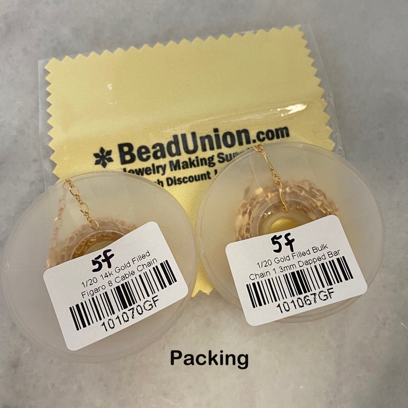 K&ouml;nnte beinhalten: Zwei Spulen mit goldgef&uuml;llter Kette, eine mit der Aufschrift "1/20 14k Gold Filled Figaro 8 Cable Chain 101070GF" und die andere mit der Aufschrift "1/20 Gold Filled Bulk Chain 1.3mm Dapped Bar 101067GF". Beide Spulen befinden sich auf einem wei&szlig;en Hintergrund mit dem Wort "Packing" darunter.