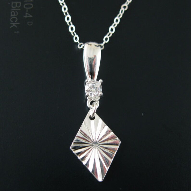 May include: A silver necklace with a diamond-shaped pendant. The pendant has a sunburst design and is accented with a small, clear stone.