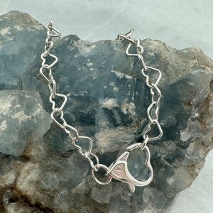 May include: A silver heart-shaped chain necklace with a heart-shaped clasp. The necklace is on a gray rock background.