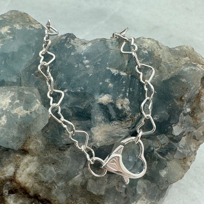 May include: A silver heart-shaped chain necklace with a heart-shaped clasp. The necklace is on a gray rock background.