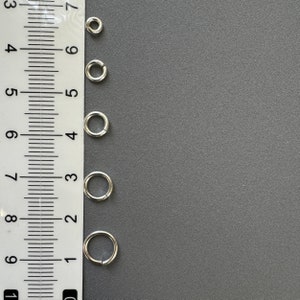May include: Five silver jump rings of different sizes, measured against a ruler. The rings are arranged vertically from largest to smallest.