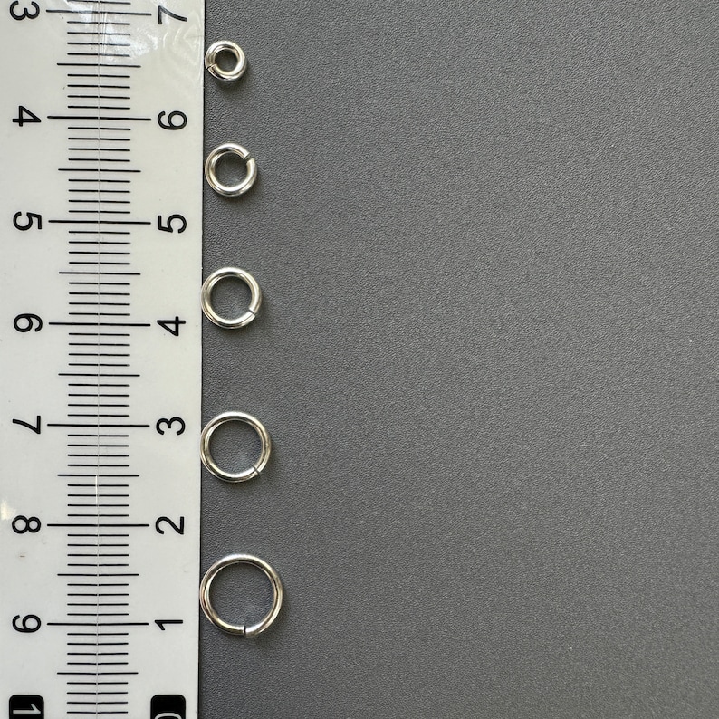 May include: Five silver jump rings of different sizes, measured against a ruler. The rings are arranged vertically from largest to smallest.