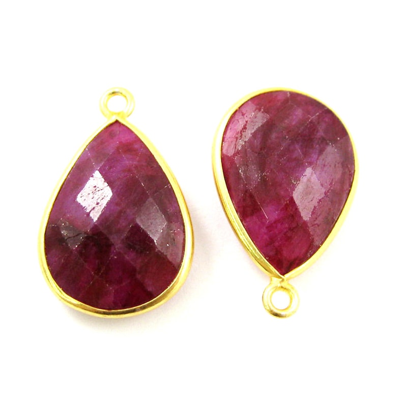 May include: Two gold-plated teardrop-shaped pendants with red ruby gemstones. The pendants have a loop at the top for attaching to a chain or cord.