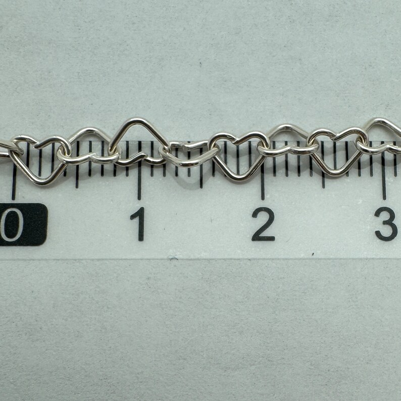 May include: Close-up of a silver heart-shaped chain measured on a ruler. The chain is approximately 2 inches long.