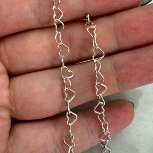 May include: Two silver heart-shaped chains, one longer than the other, are shown against a light brown background.