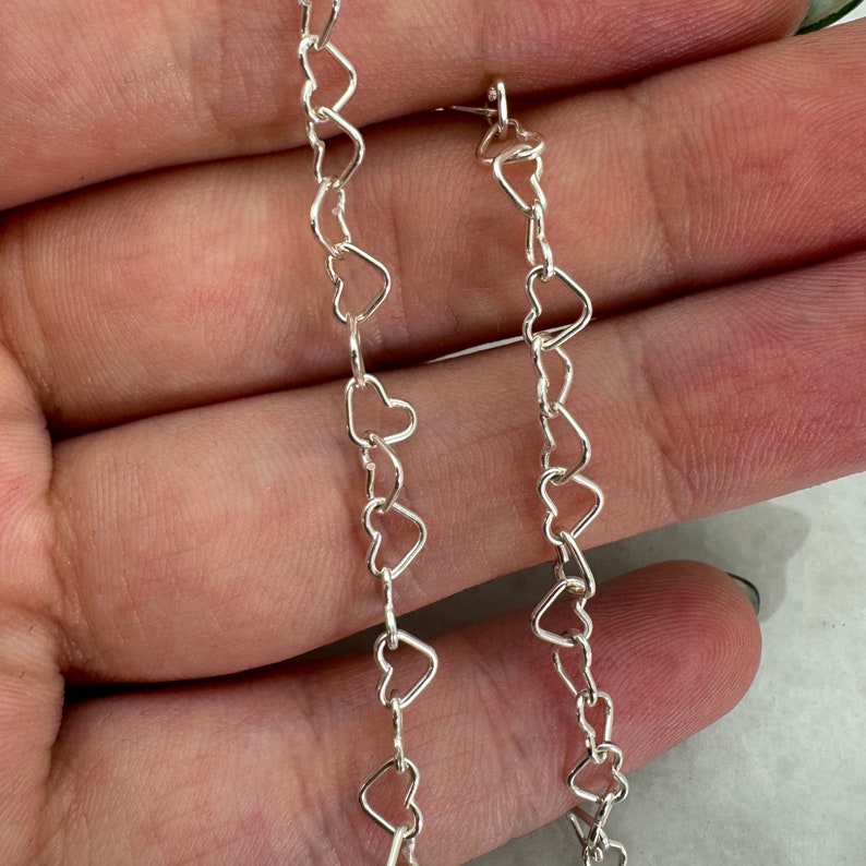 May include: Two silver heart-shaped chains, one longer than the other, are shown against a light brown background.