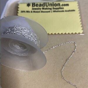 May include: A spool of silver chain with a small section of the chain laid out next to it. The spool is clear plastic and the chain is silver in colour. The text on the yellow label reads "BeadUnion.com Jewelry Making Supplies 50% Mix & Match Discount | Wholesale Available".