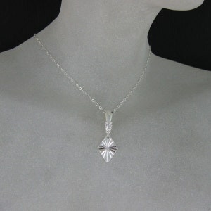 May include: A silver necklace with a diamond-shaped pendant. The pendant has a textured, sunburst design.