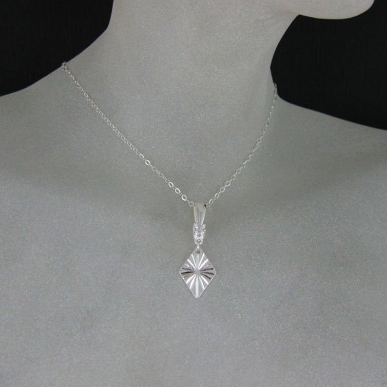 May include: A silver necklace with a diamond-shaped pendant. The pendant has a textured, sunburst design.