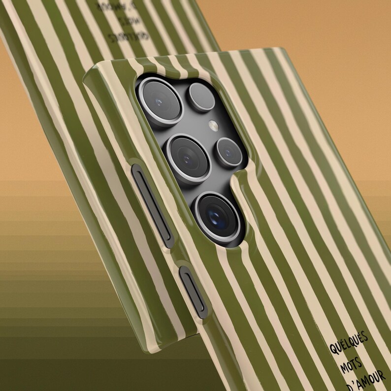 May include: A close-up of two phone cases with a green and beige striped pattern. The cases have multiple camera lenses and the text "QUELQUES MOTS D'AMOUR" is visible on one case. The cases are angled towards the viewer.