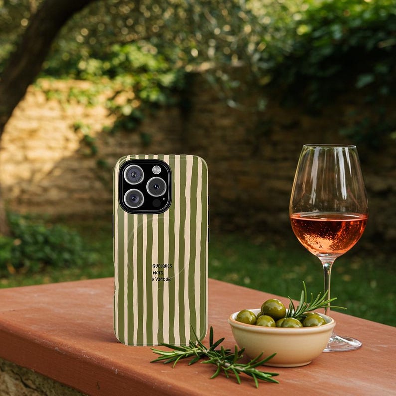 May include: A green and cream striped phone case with the text "QUELQUES MOTS D'AMOUR" is displayed on a red-brown surface. A glass of rose wine, a small bowl of olives, and rosemary sprigs are also present, suggesting a relaxed outdoor setting.