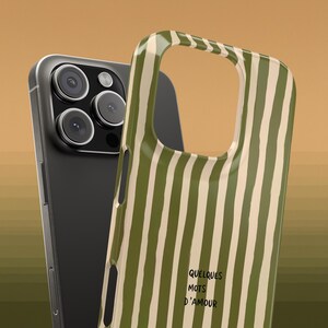 May include: A black smartphone with a triple-lens camera is next to a green and white striped phone case. The case has the words "QUELQUES MOTS D'AMOUR" printed on it. The background is a solid tan color.