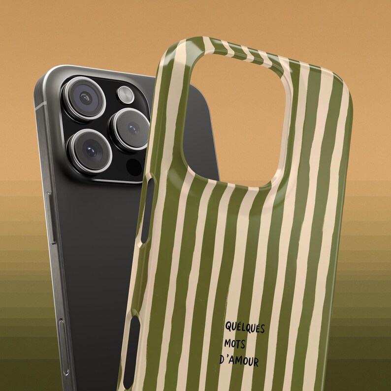 May include: A black smartphone with a triple-lens camera is next to a green and white striped phone case. The case has the words "QUELQUES MOTS D'AMOUR" printed on it. The background is a solid tan color.