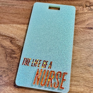 May include: A rectangular, light blue badge holder with glitter. The text "THE LIFE OF A NURSE" is in orange and red glitter. The badge holder has a slot at the top for a lanyard or clip.