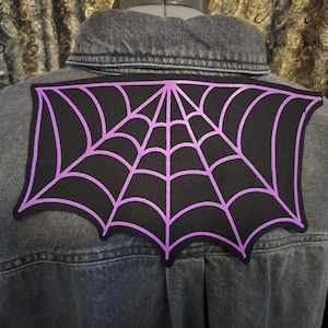 May include: A black patch featuring a purple spiderweb design is attached to the back of a denim jacket. The spiderweb is a detailed graphic, with a dark background and a contrasting purple outline. The jacket is a faded grey colour.