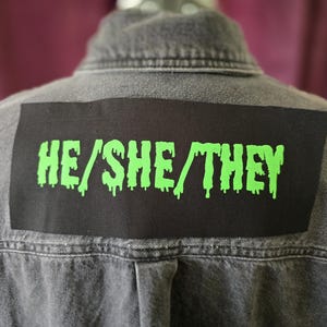 May include: A gray denim jacket with a black patch on the back. The patch features the text "HE/SHE/THEY" in dripping neon green letters. The jacket has a classic collar and button-down front.