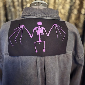 May include: A grey denim jacket with a black patch featuring a pink skeleton bat design. The jacket is displayed on a mannequin, showcasing the back of the garment. The design is centered on the upper back.